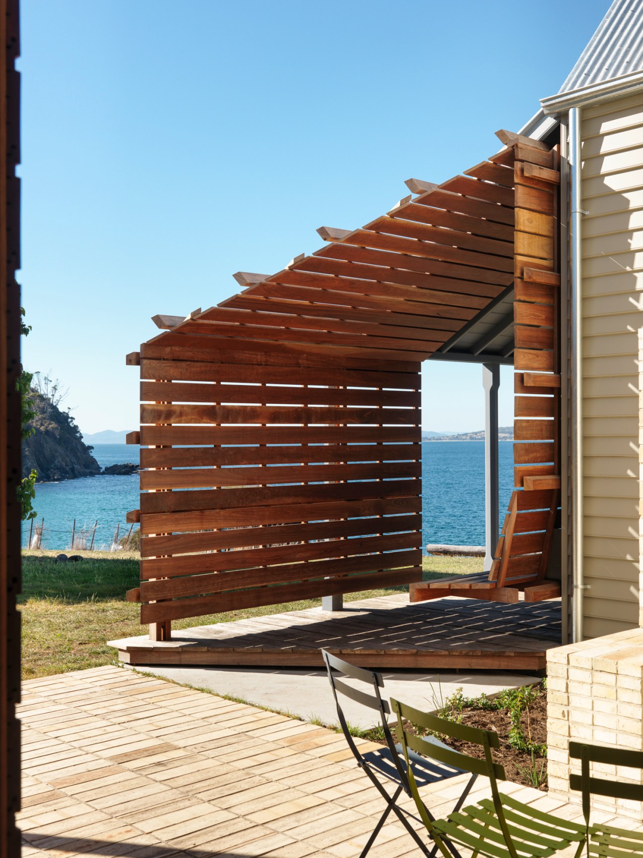 Wooden pergola structure with slatted privacy screen attached to white weatherboard house overlooking water
