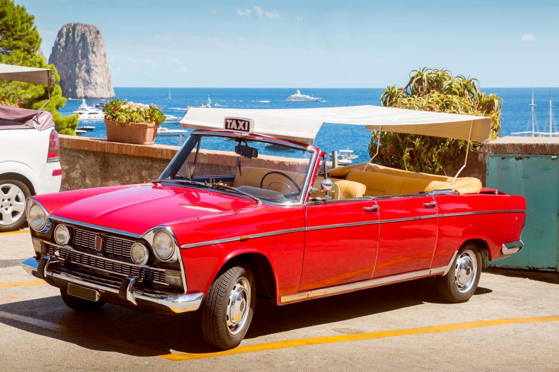 Red convertible taxi parked on Capri with scenic coastal views and Faraglioni rock formations in background