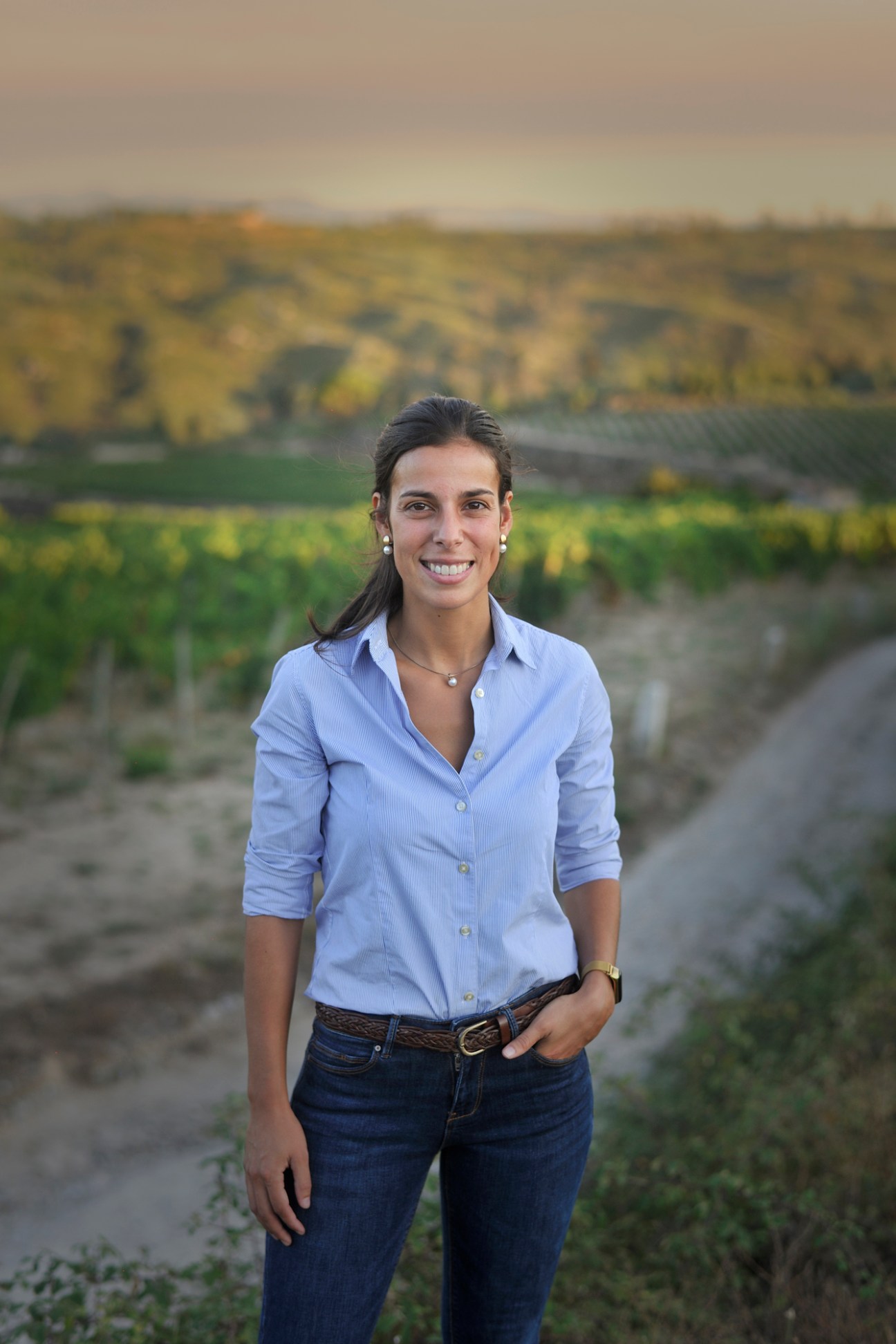 Mafalda Perdigao standing in vineyard rows wearing light blue shirt and jeans with hills in background