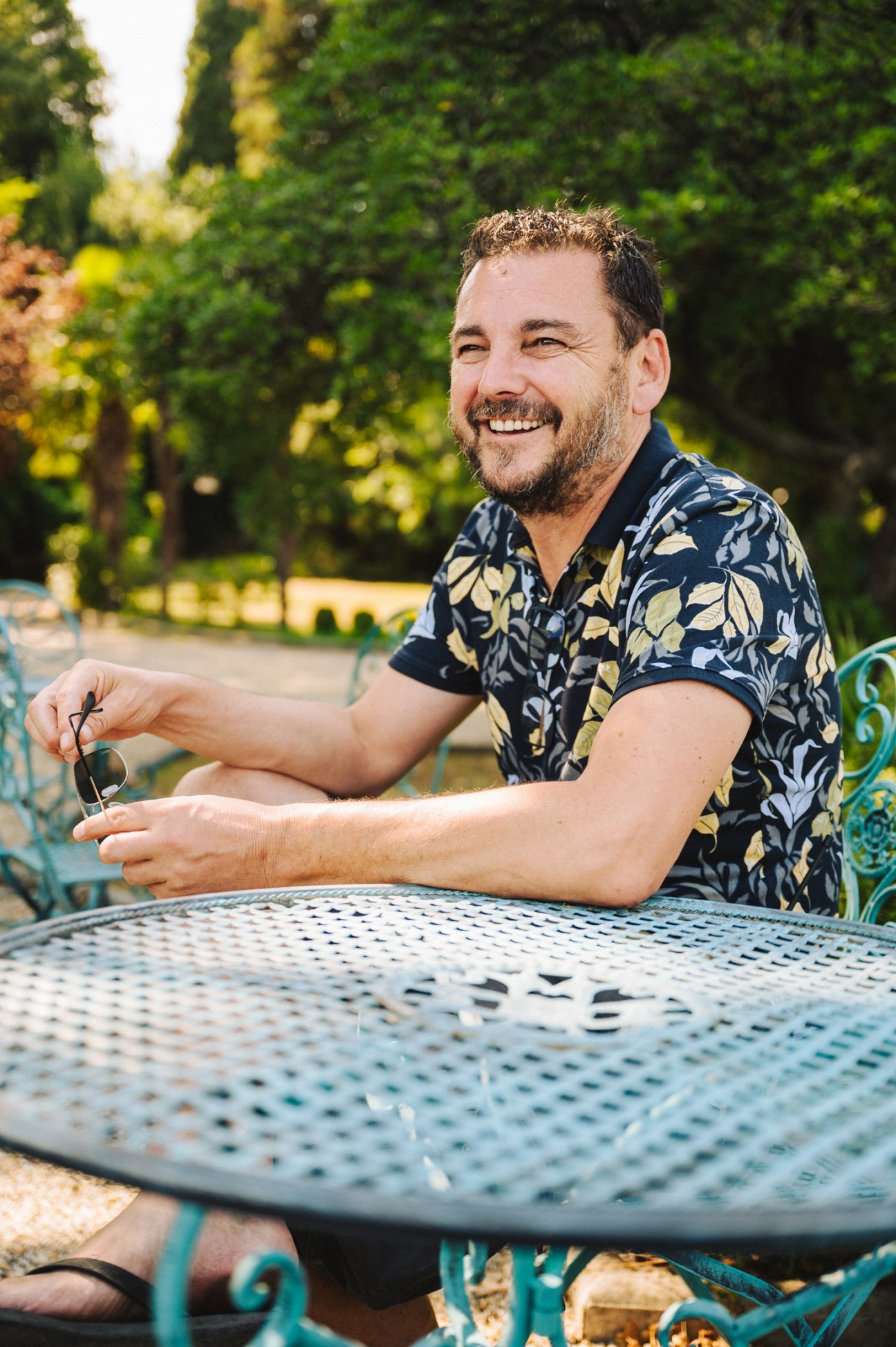 Dean Lalic smiling while sitting at an outdoor table holding sunglasses
