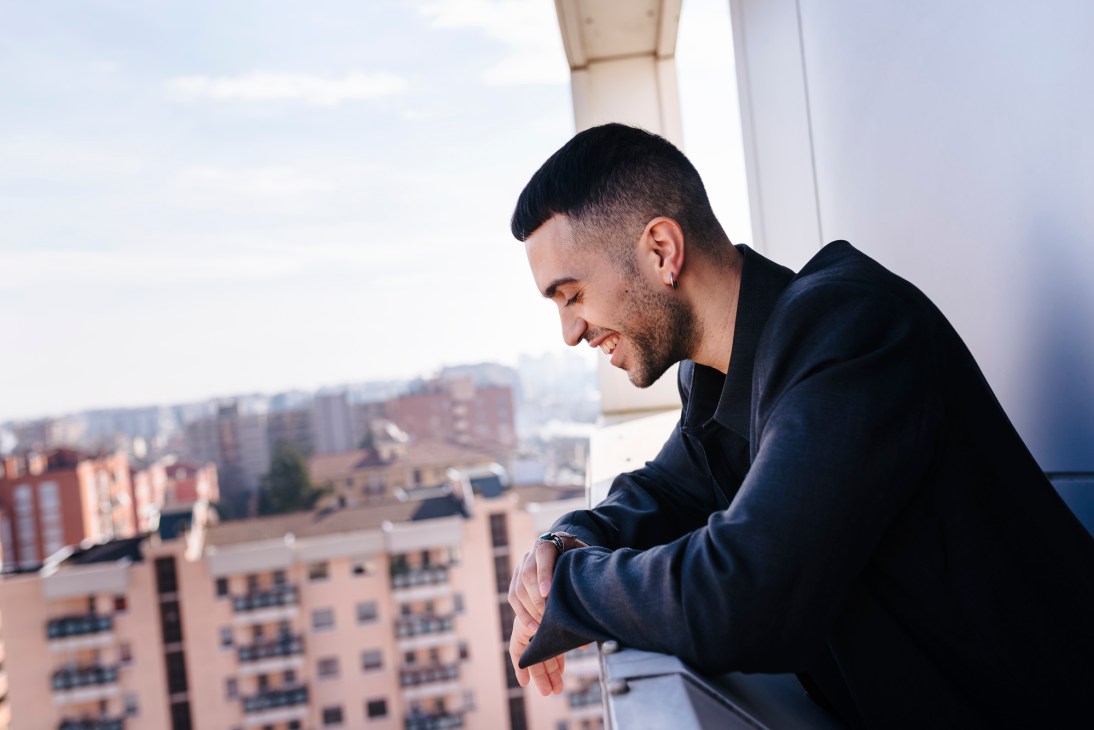 Young man in black hoodie smiling while looking down from a balcony with urban buildings in background