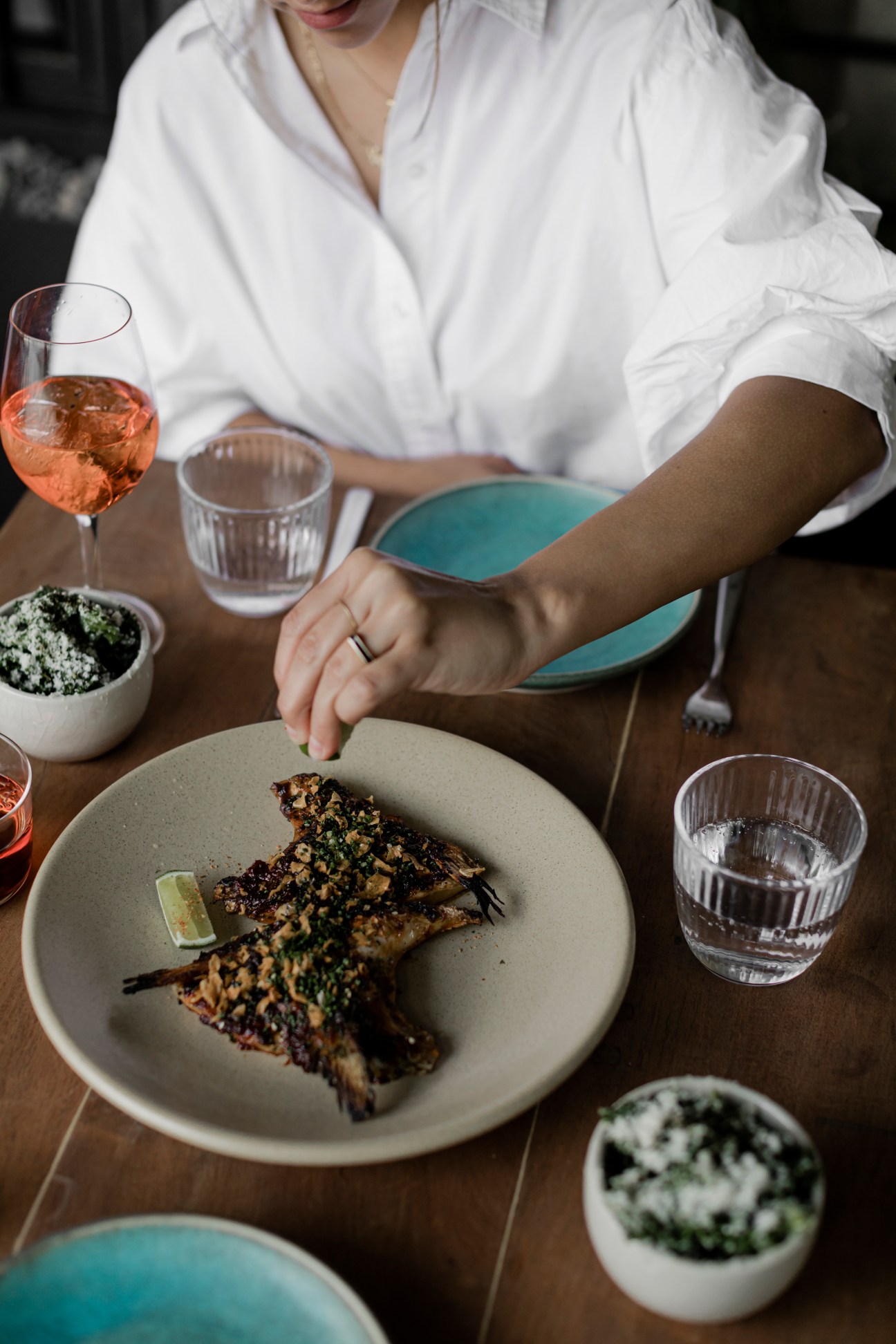 Person in white shirt dining on grilled fish collars with herbs, rosé wine, and side dishes at EM restaurant.