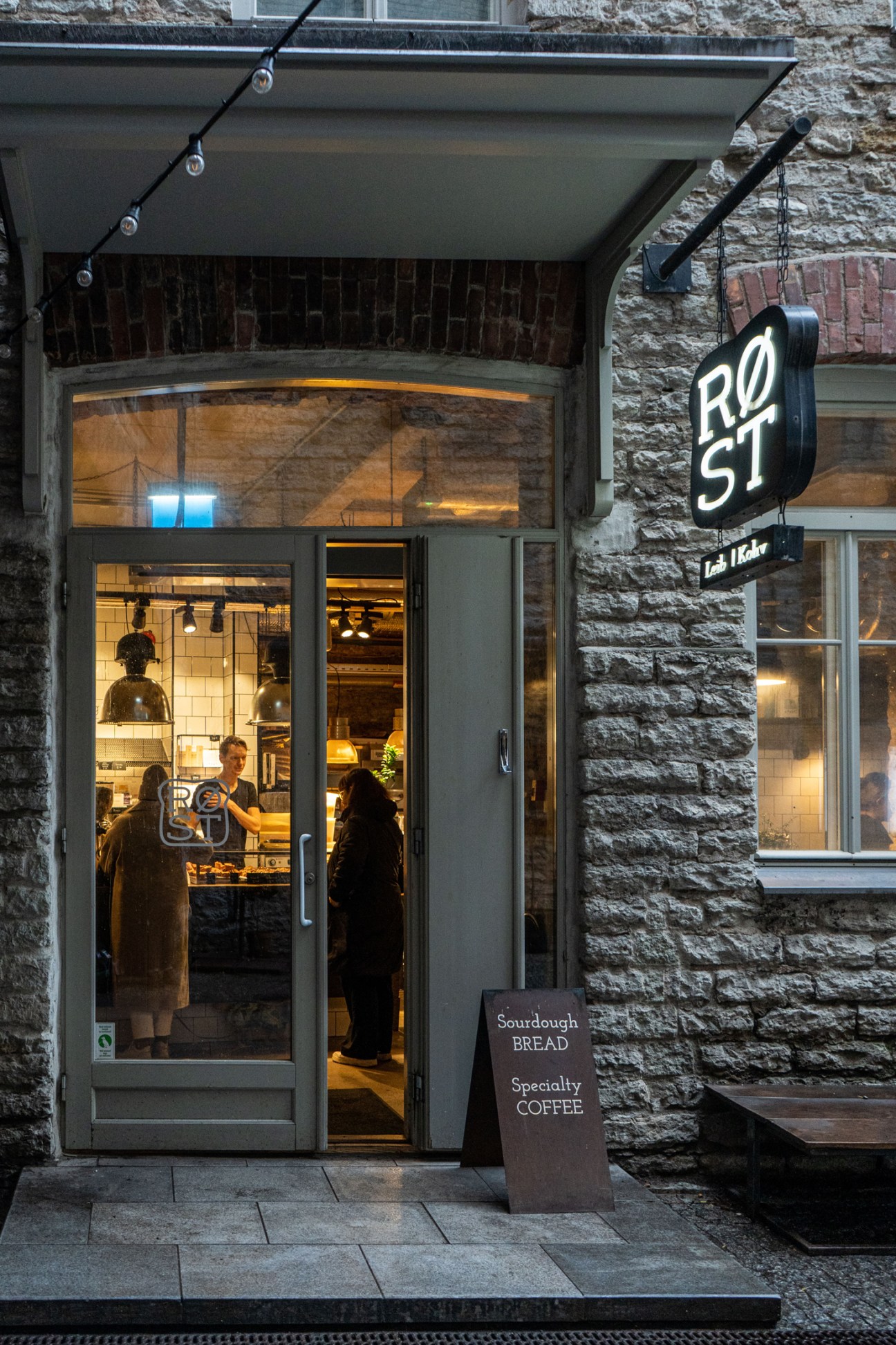 Exterior of Røst bakery with stone walls, glass storefront, and chalkboard sign advertising sourdough bread and specialty coffee