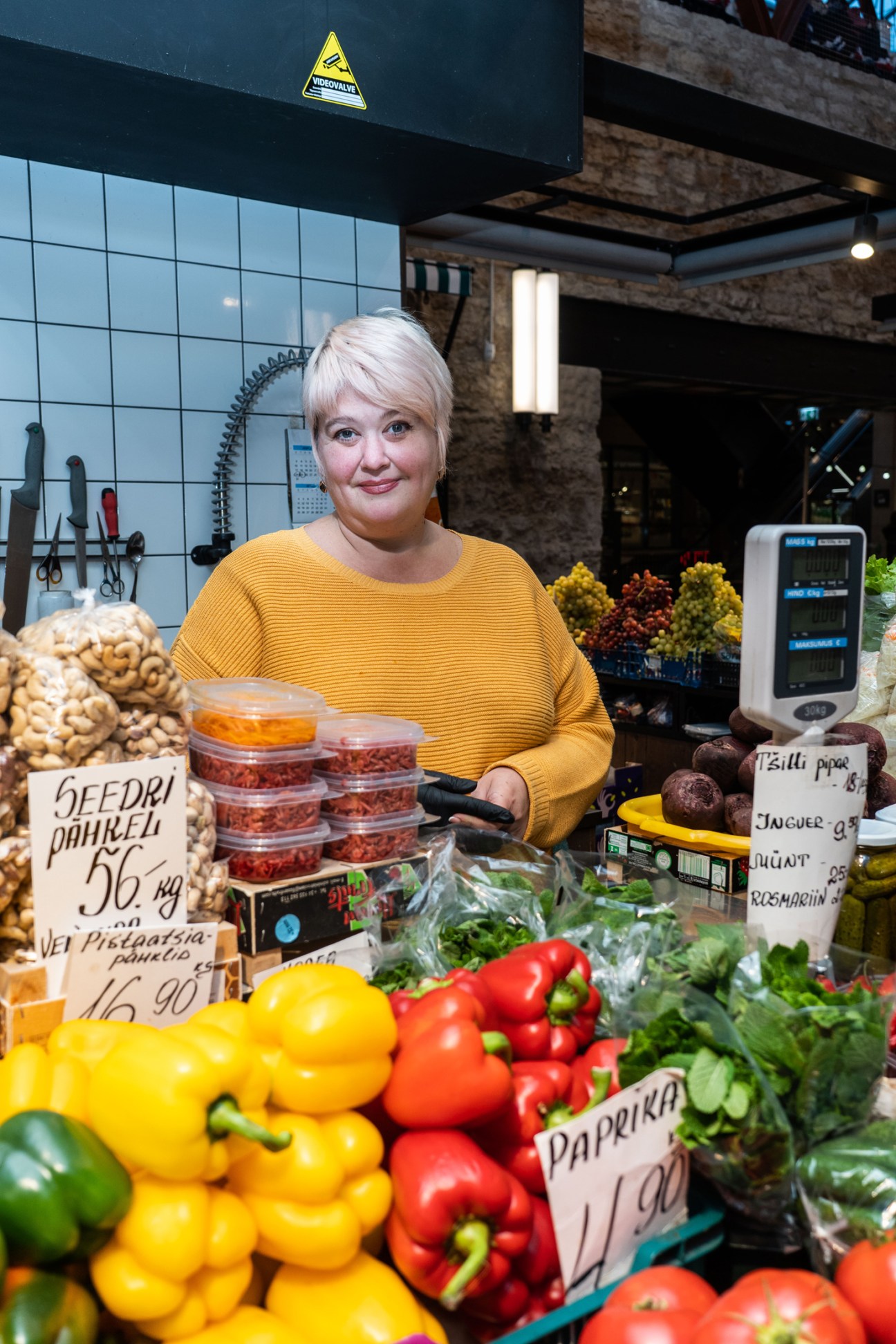 Woman in yellow sweater at market stall with colorful peppers and produce with price signs