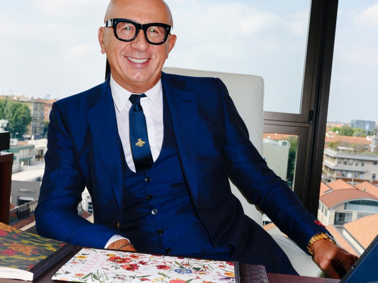 Marco Bizzarri sits at his office desk wearing a blue three-piece suit and glasses, smiling at the camera