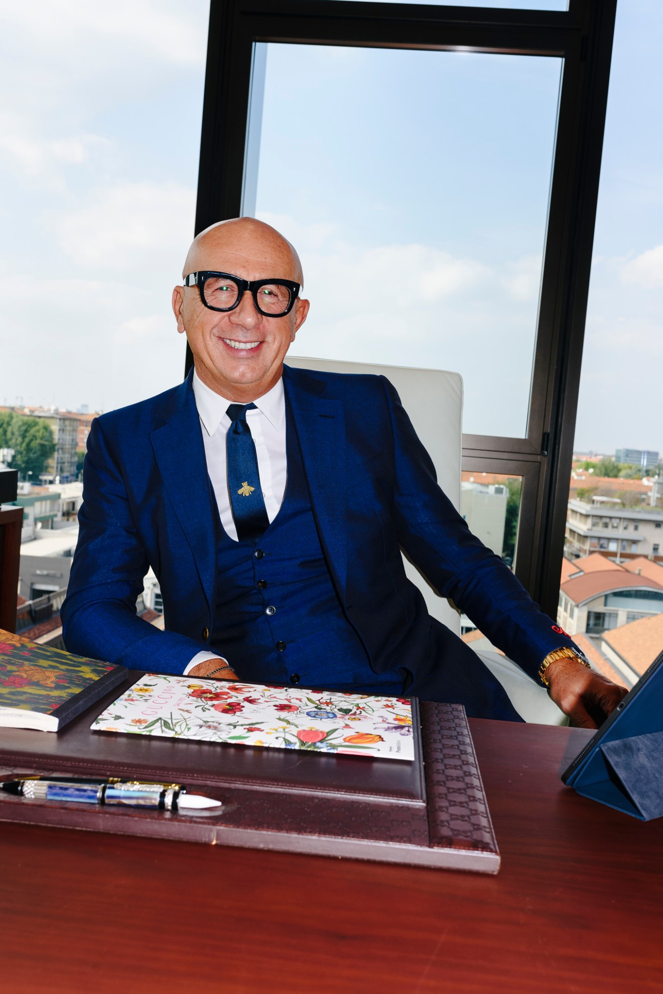 Marco Bizzarri sits at his office desk wearing a blue three-piece suit and glasses, smiling at the camera