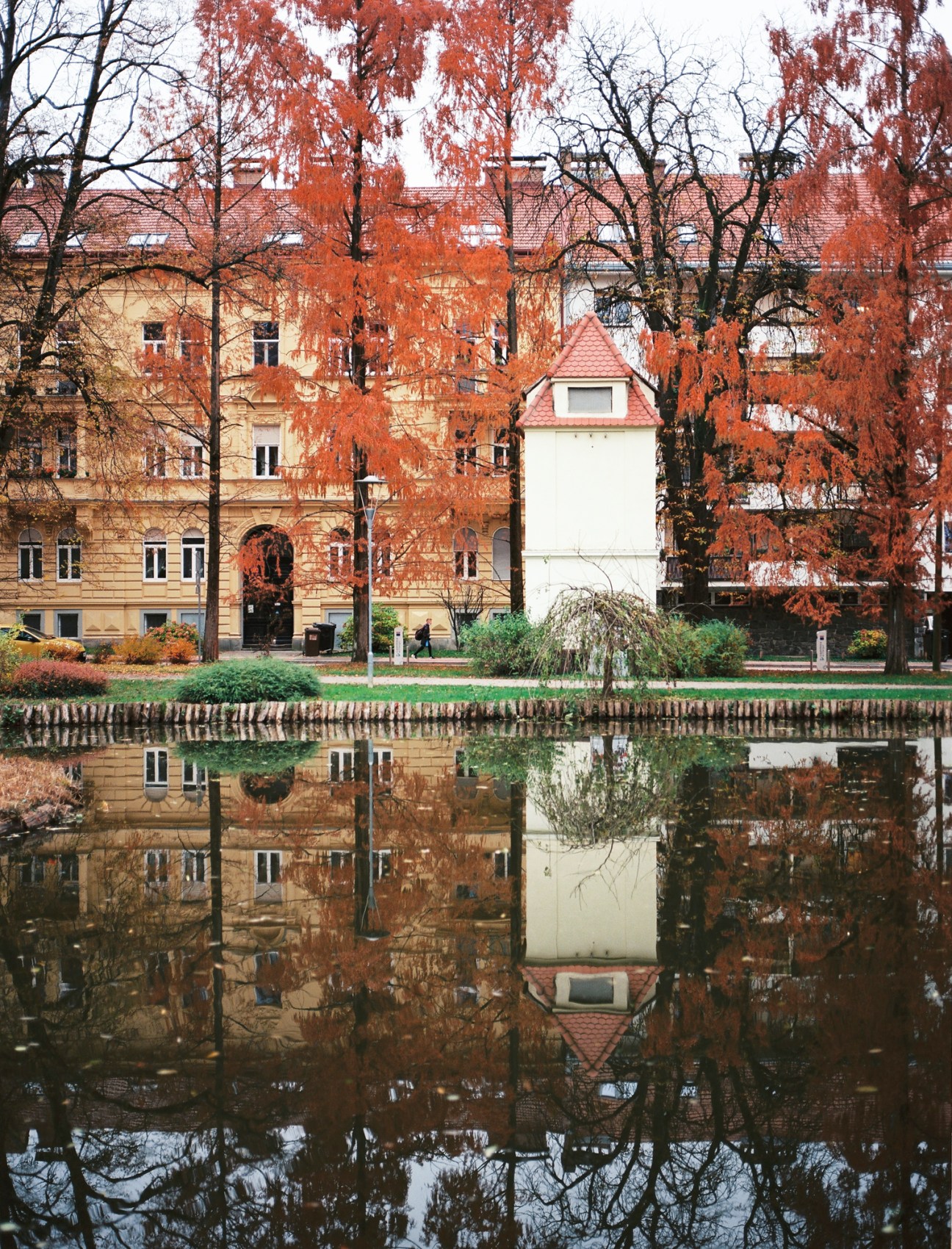 Autumn scene in Maribor with orange-leafed trees reflected in calm river water, historic buildings visible in background