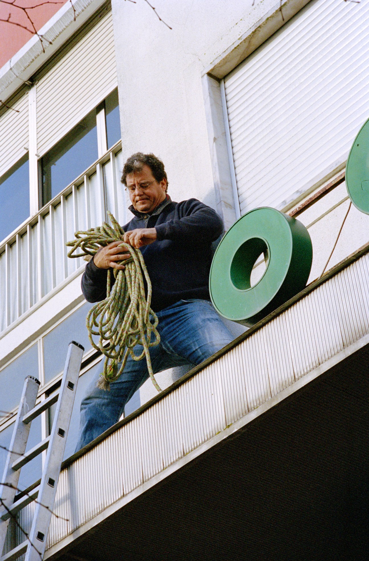 Man on building balcony handling rope while removing large green neon sign letters