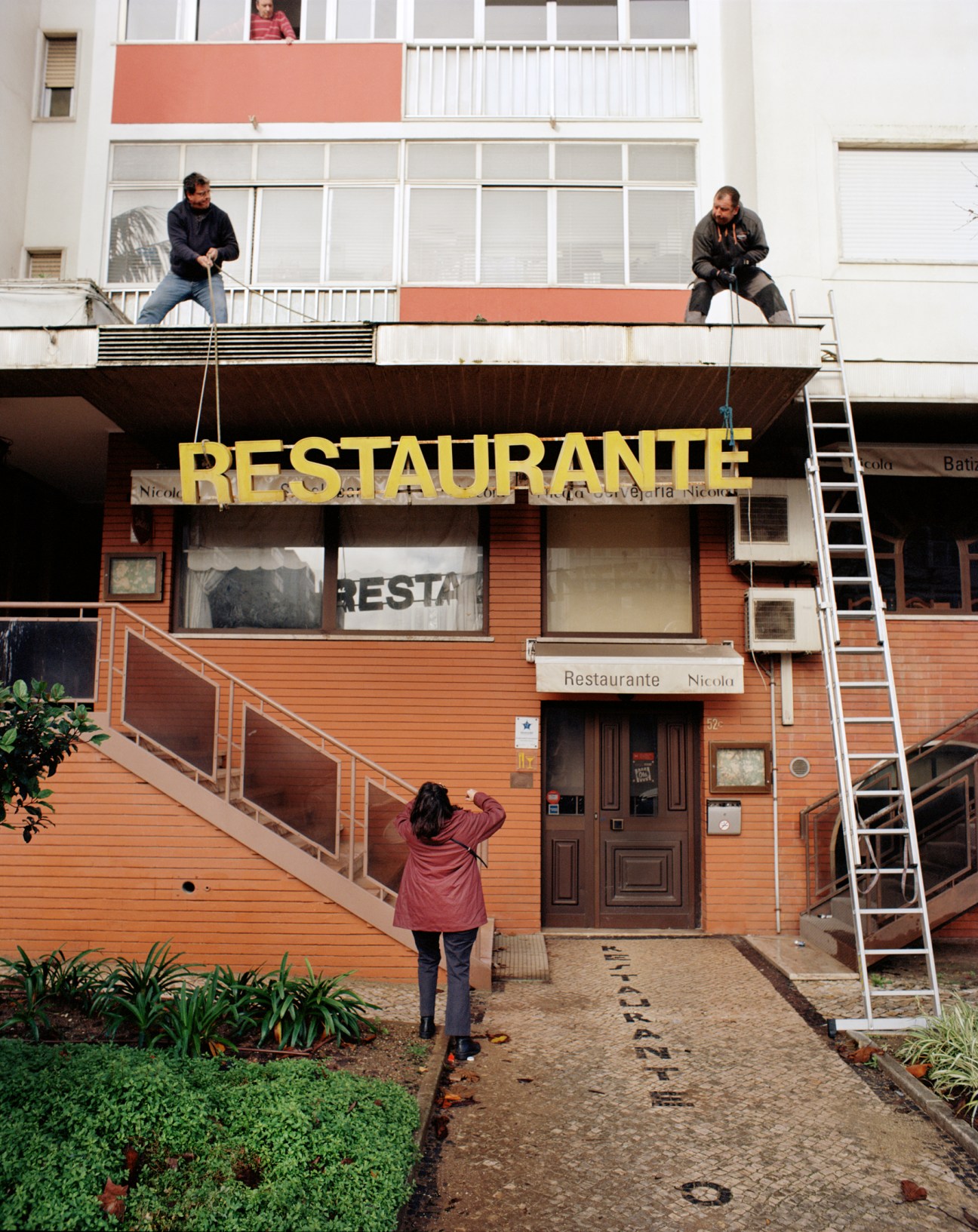 Two workers carefully lowering a large yellow 'RESTAURANTE' neon sign from a building balcony using ropes
