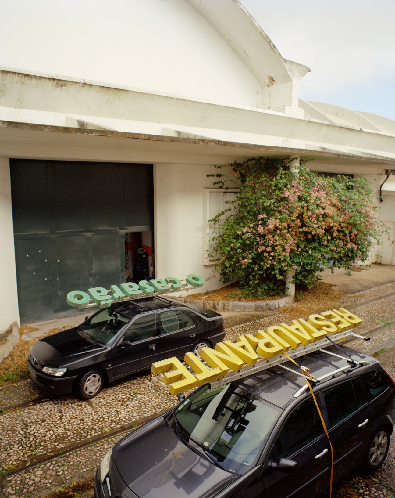 Green metal letters spelling O CASARÃO strapped to the roof of a dark Renault car parked outside a white building