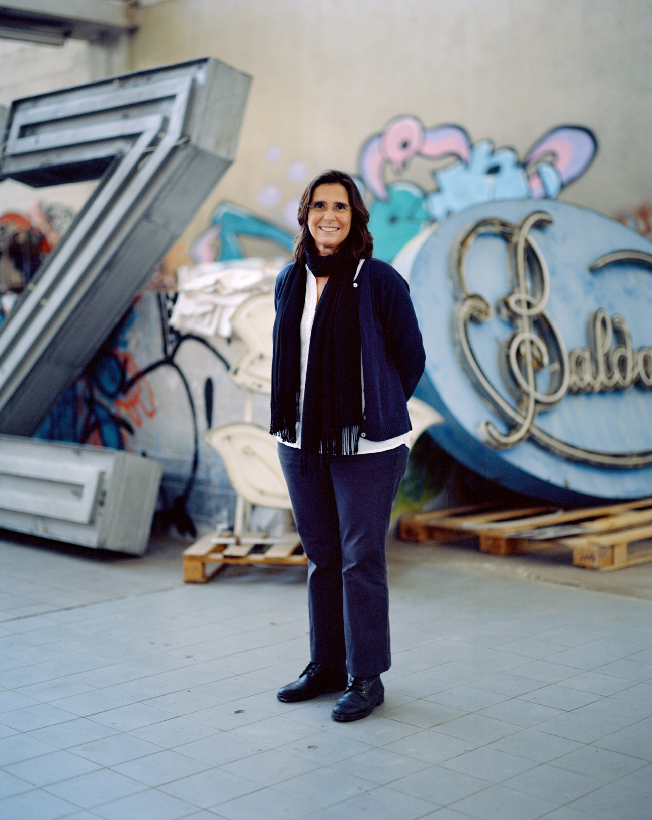 Rita Múrias stands smiling in a warehouse space with colorful vintage neon signs and lettering visible in the background