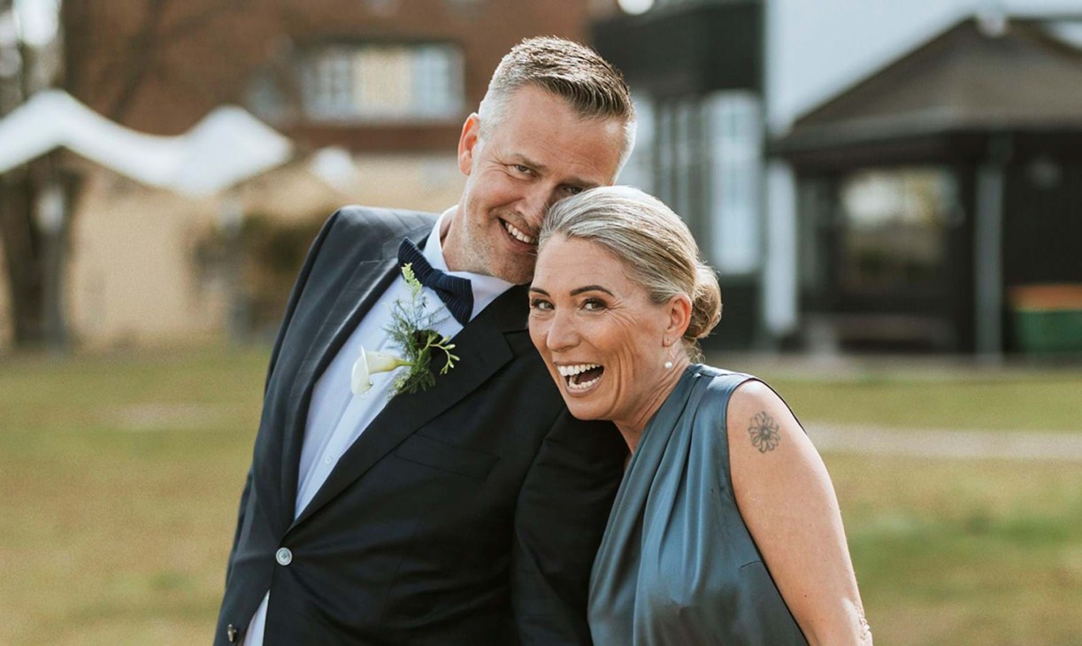 A smiling couple pose together outdoors, the man in a dark suit with a flower boutonniere and the woman in a sleeveless blue-grey dress.