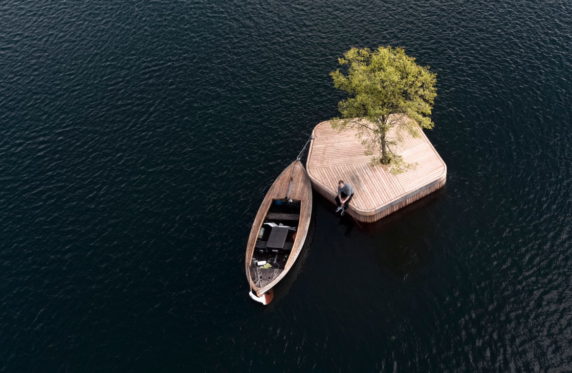 Small floating wooden platform with tree in Copenhagen harbor with person sitting and boat moored alongside