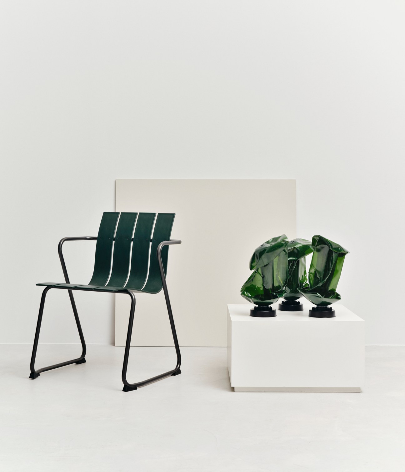 Green chair with vertical slats and metal frame next to white pedestal with three large emerald crystal objects