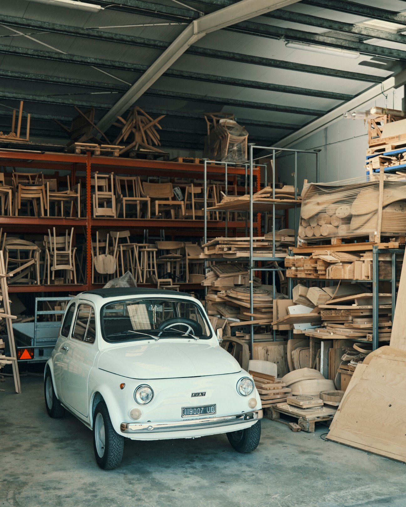 White vintage Fiat 500 parked inside Mattiazzi furniture factory warehouse surrounded by wooden chairs and materials