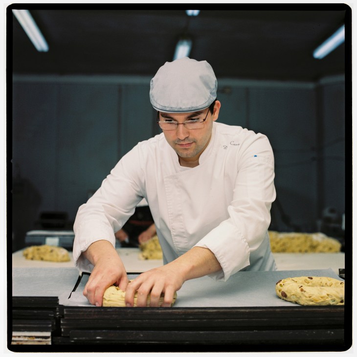 Baker in white chef's coat and cap working with dough on a counter in a professional kitchen.