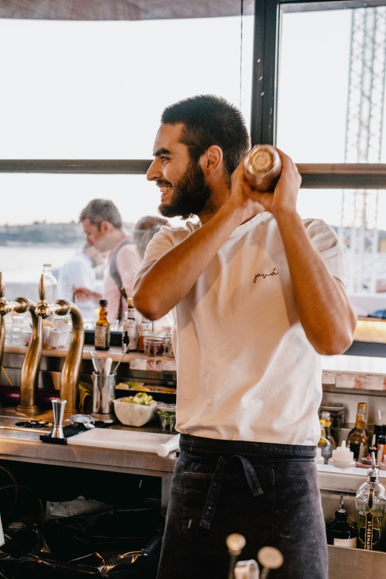 Bartender in white shirt shaking cocktail behind bar counter with bottles and glassware