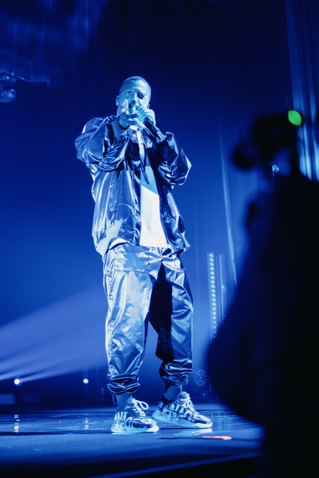 Musician Dino d'Santiago performing on stage in a shiny black outfit while singing into a microphone under blue stage lighting.