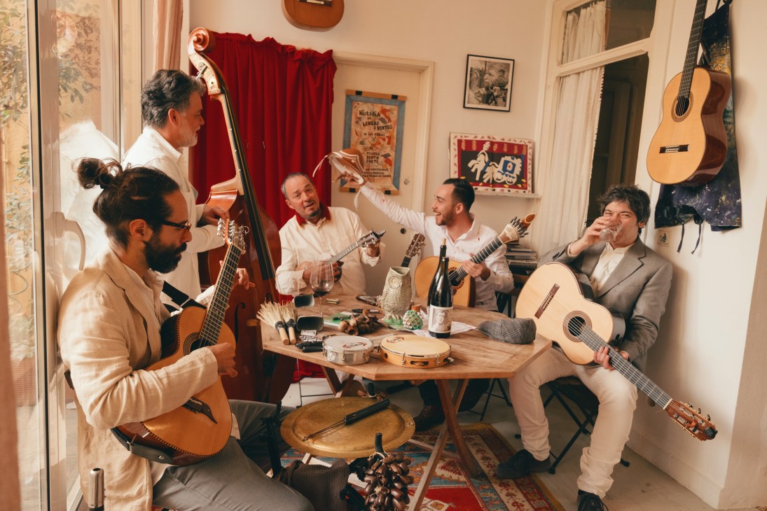 Musicians playing acoustic guitars and traditional instruments during a jam session in a light-filled living room.