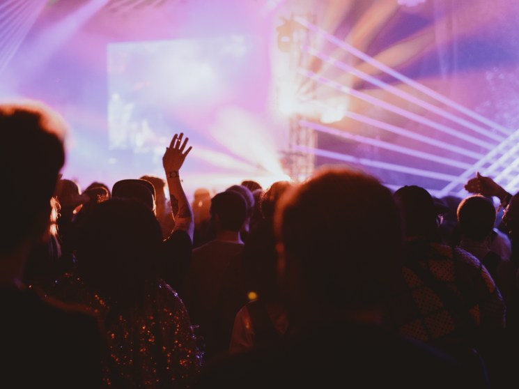 Silhouetted concert crowd with raised hands against dramatic purple and orange stage lighting at music festival