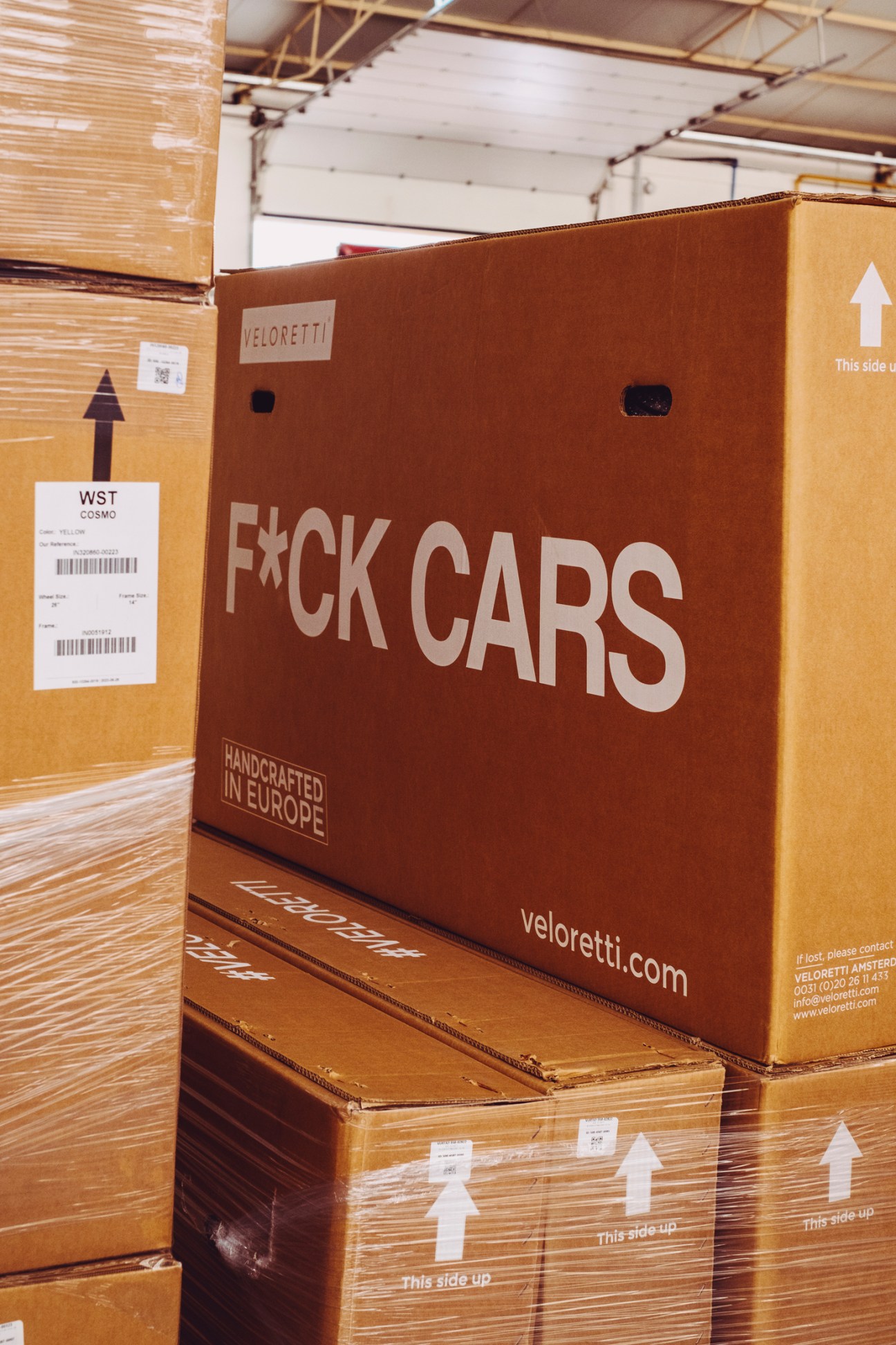 Large brown cardboard box with 'F*CK CARS' slogan and Veloretti branding in a warehouse with stacked packages