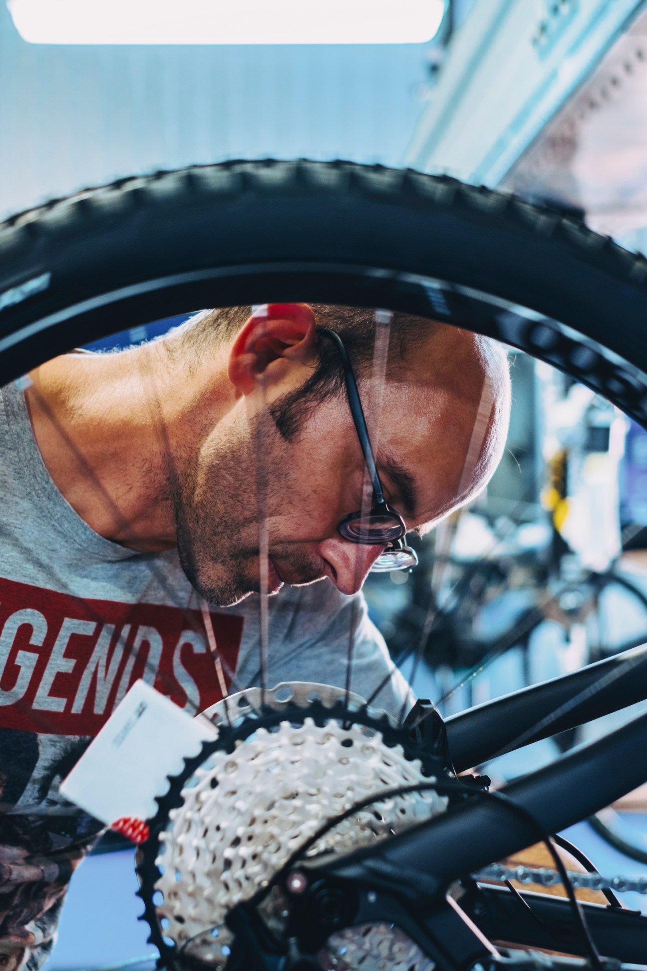 A bicycle mechanic wearing glasses inspects a bike wheel and drivetrain components in a factory setting