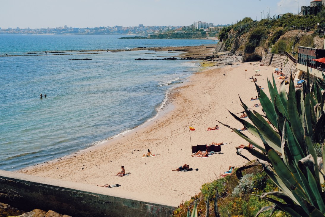 Beachgoers relaxing on a sandy cove below clifftop buildings with Mediterranean coastline in the distance