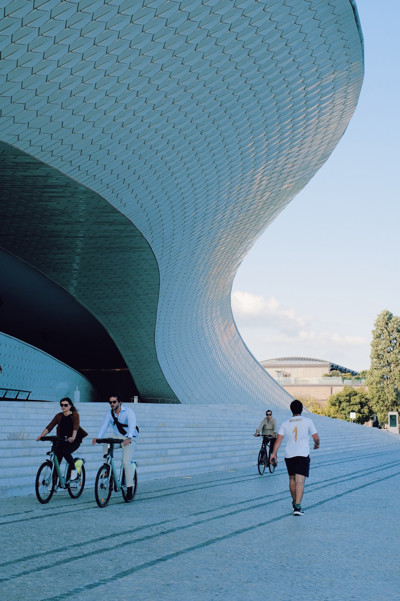 People cycling and jogging in front of a modern curved turquoise building with patterned brick facade
