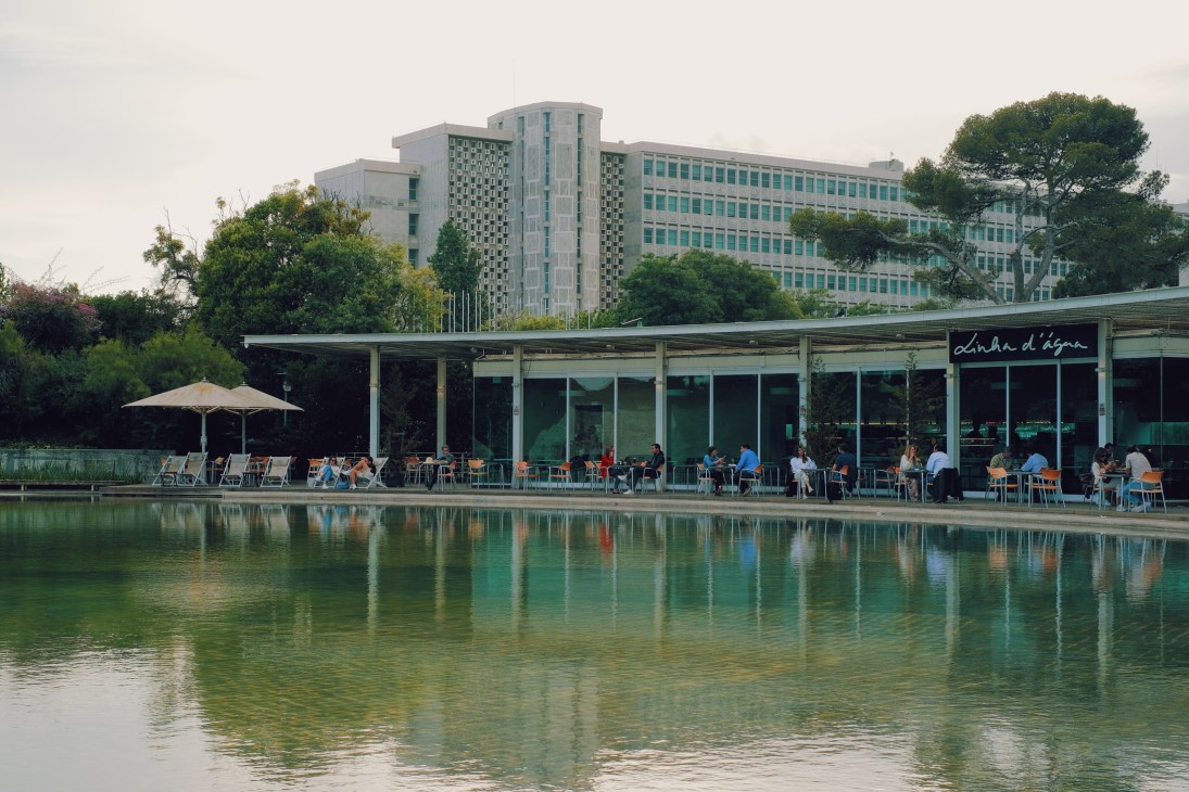 Waterfront café with diners seated along a canal, modern mid-rise buildings in background, trees and umbrellas visible