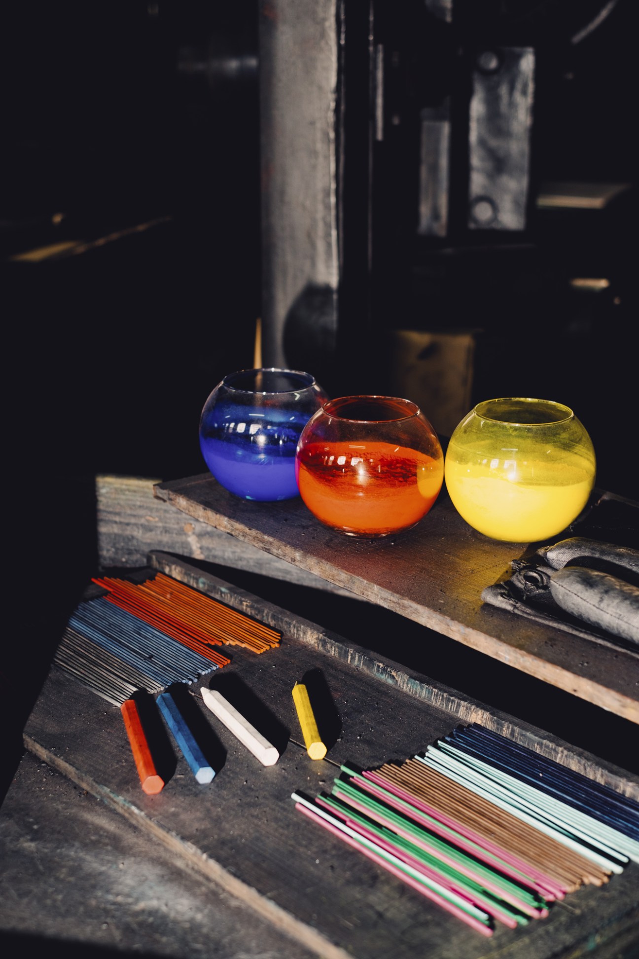 Three colorful glass bowls containing blue, red, and yellow materials next to multicolored pencil rods on wooden surface