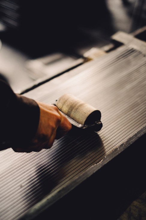 Close-up of a hand using a plane tool to smooth ridged wooden pencil slats in a workshop