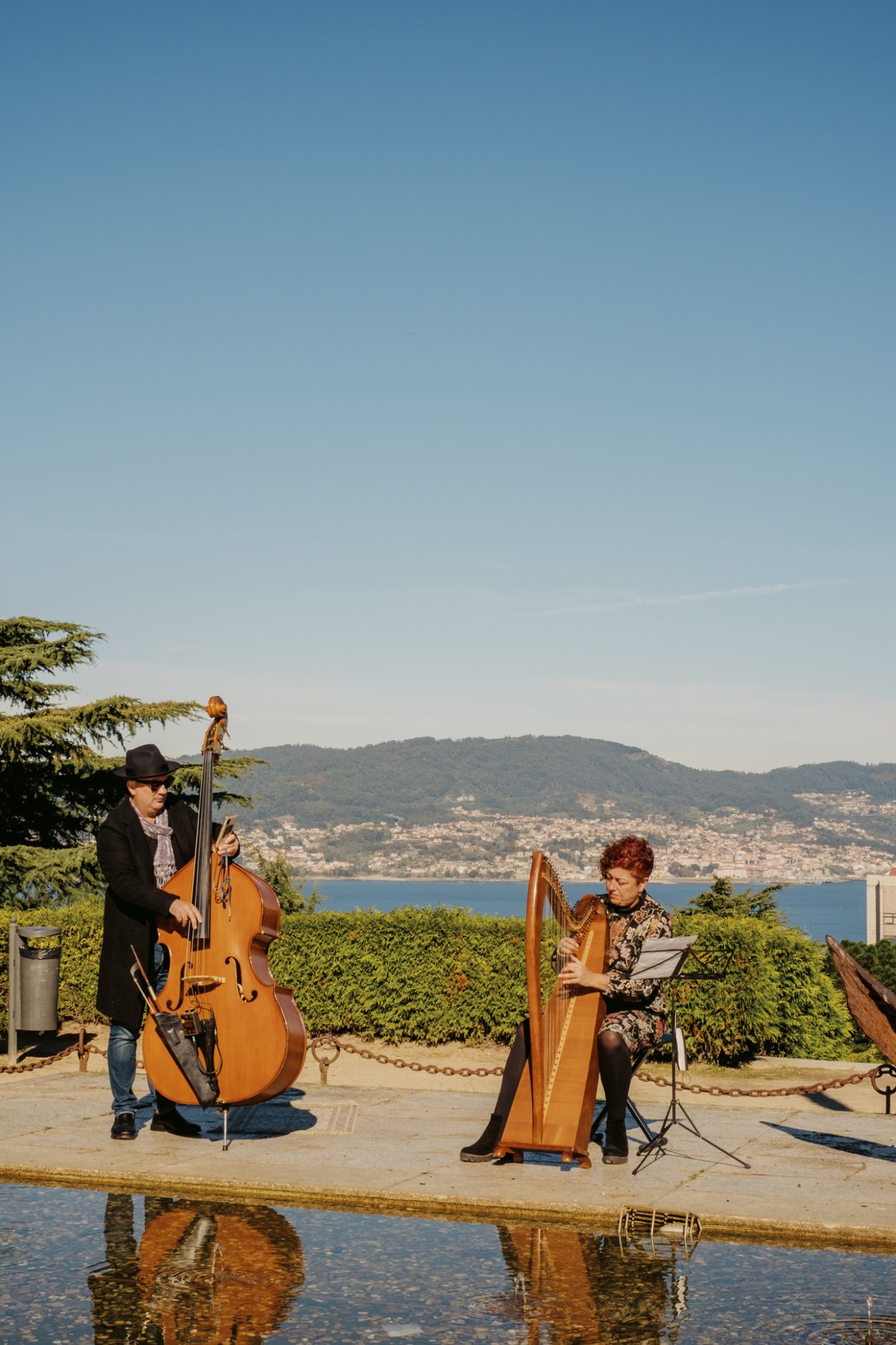 Two street musicians performing outdoors with upright bass and harp overlooking Vigo's estuary and hills