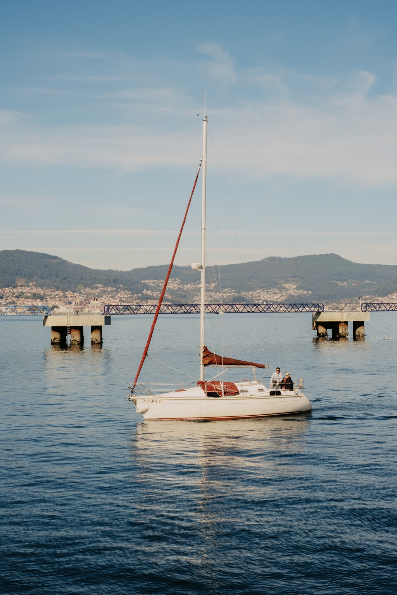White sailboat with red sail on calm blue waters with bridge and hills in background