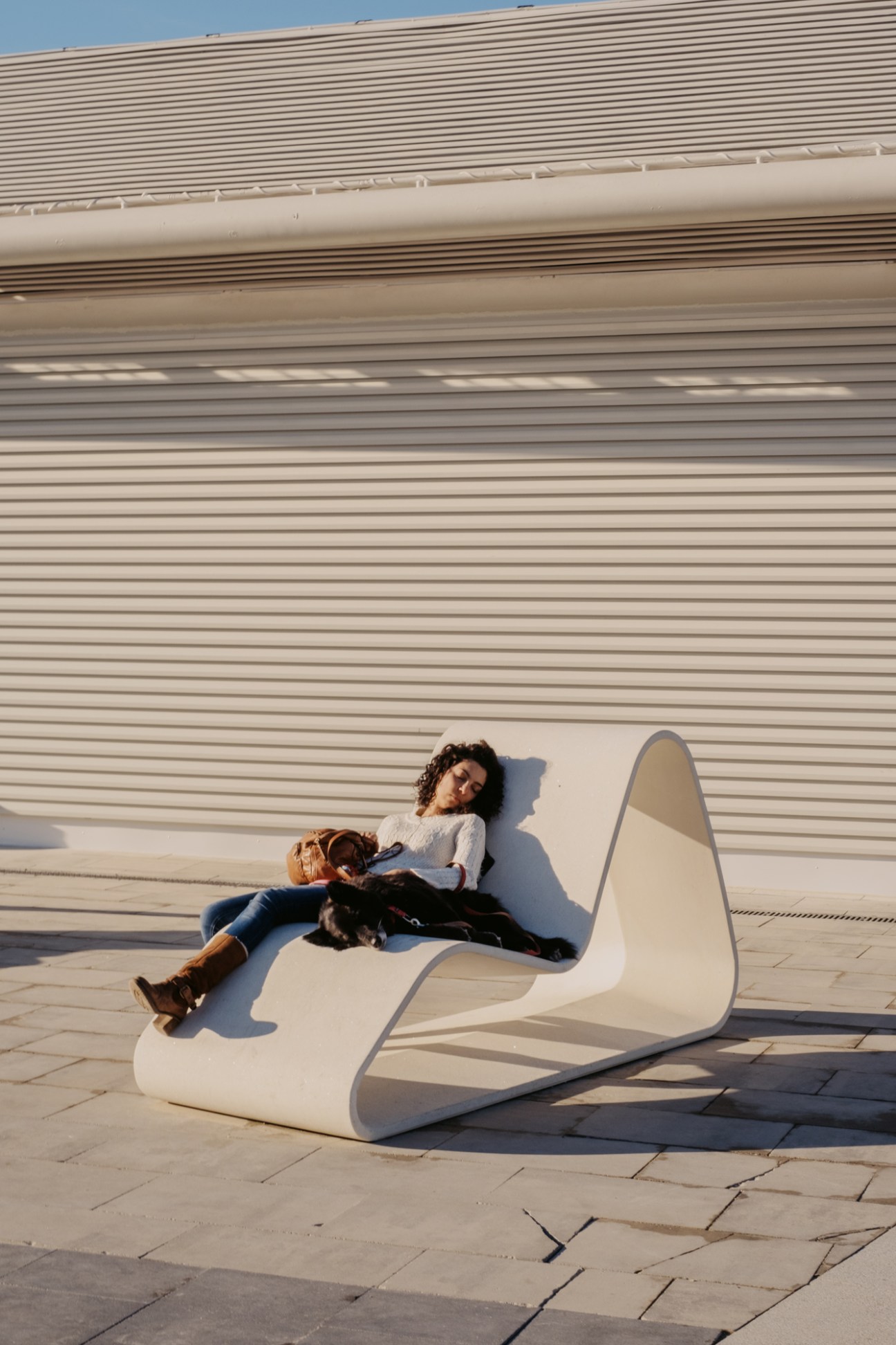 Woman with curly hair relaxing on modern white sculptural bench in sunny outdoor setting with dog