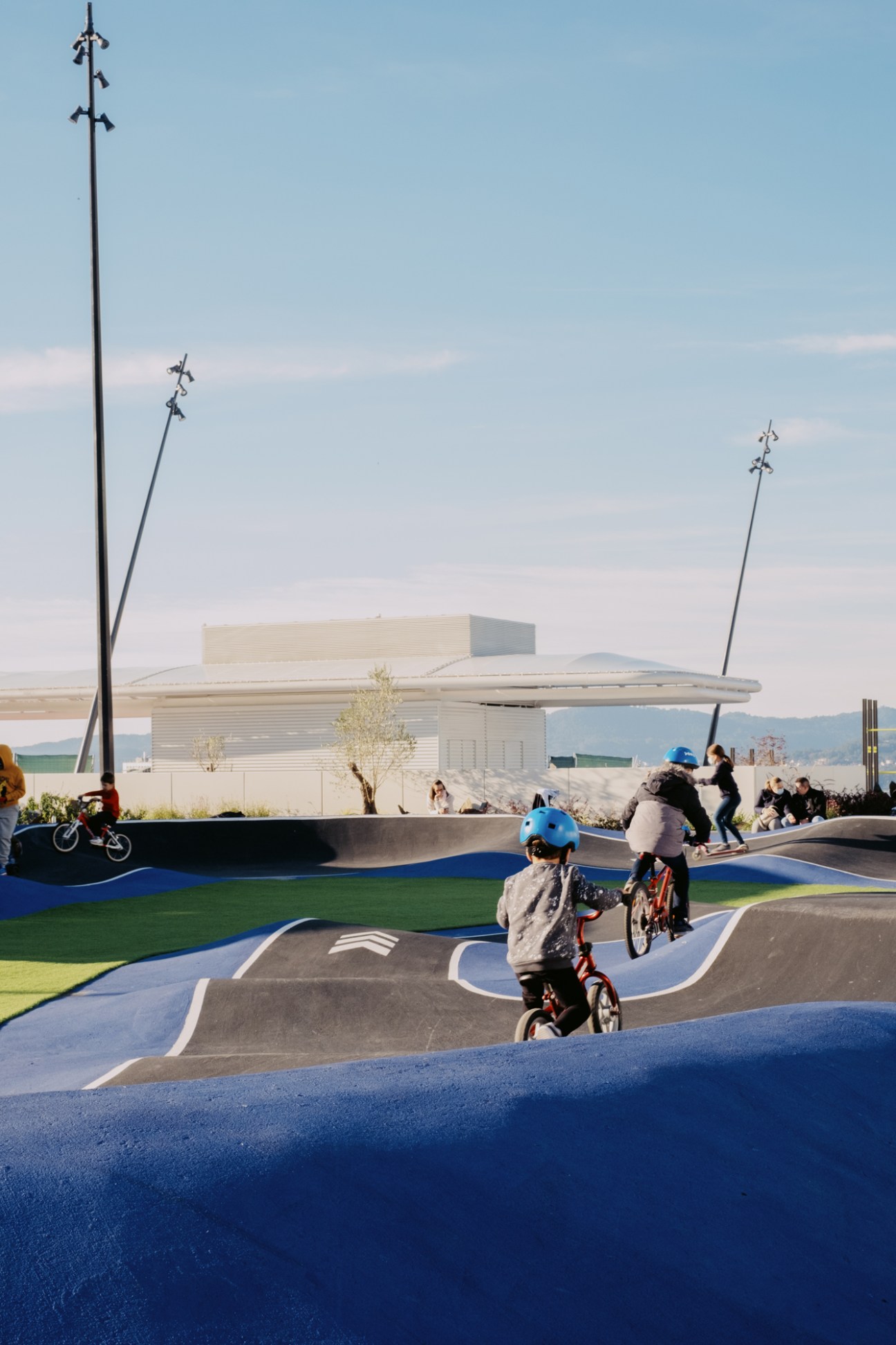 Children riding bikes on colorful curved track at recreational rooftop facility with modern building in background