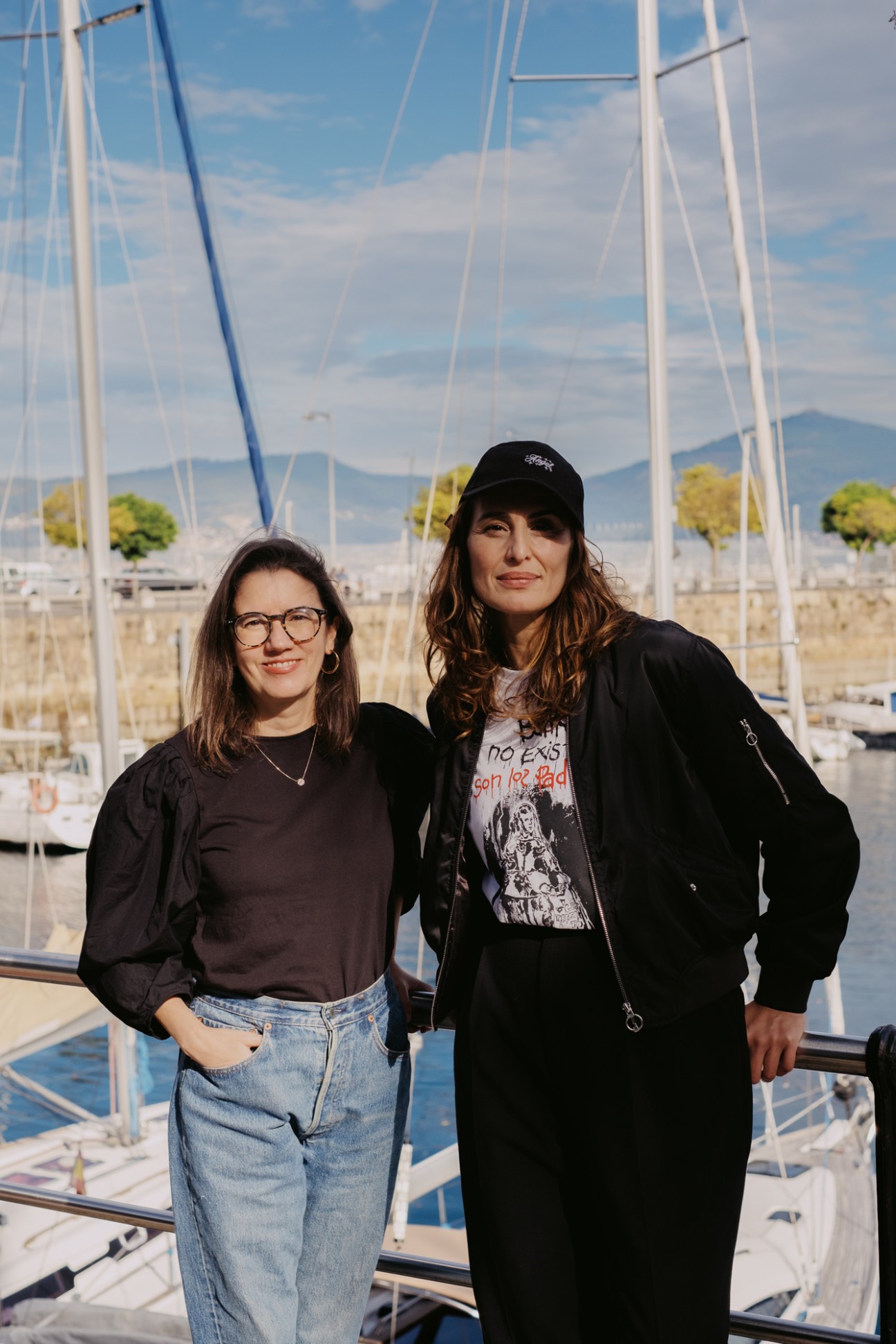 Two women from O Cable Inglés standing together outdoors in a marina with sailboat masts in the background