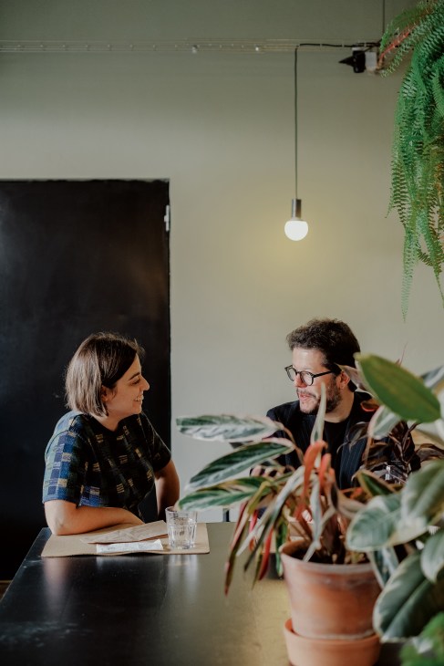 Two people in conversation at a desk in a modern office space with potted plants and hanging greenery.