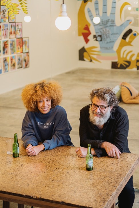 Two people sit at a cork table with beer bottles in a gallery space with colorful artwork on walls