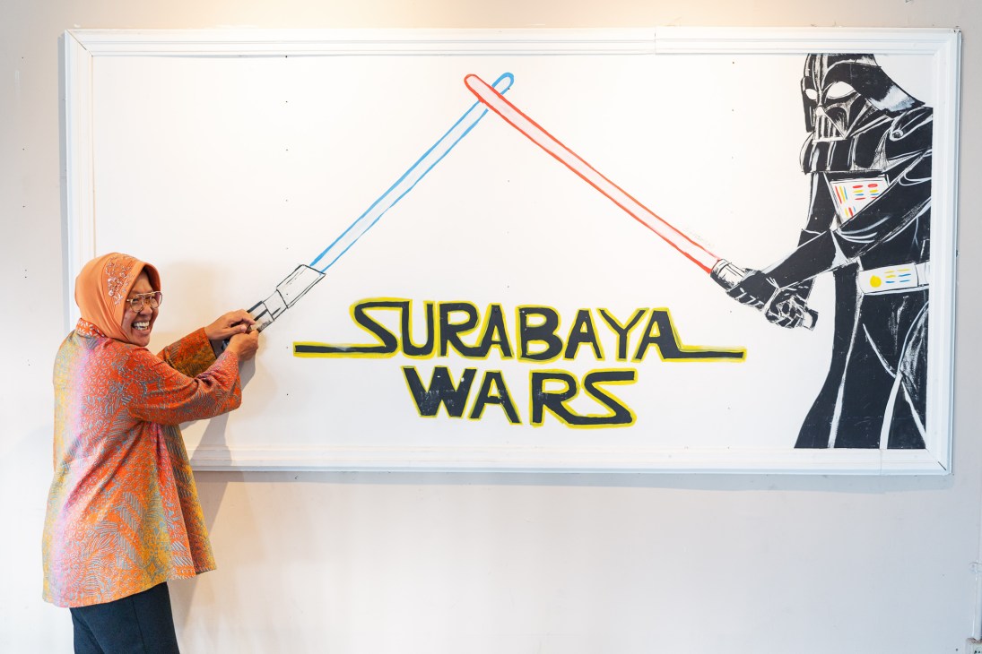 Woman in orange headscarf and colorful clothing holding lightsaber prop next to Surabaya Wars poster with Darth Vader
