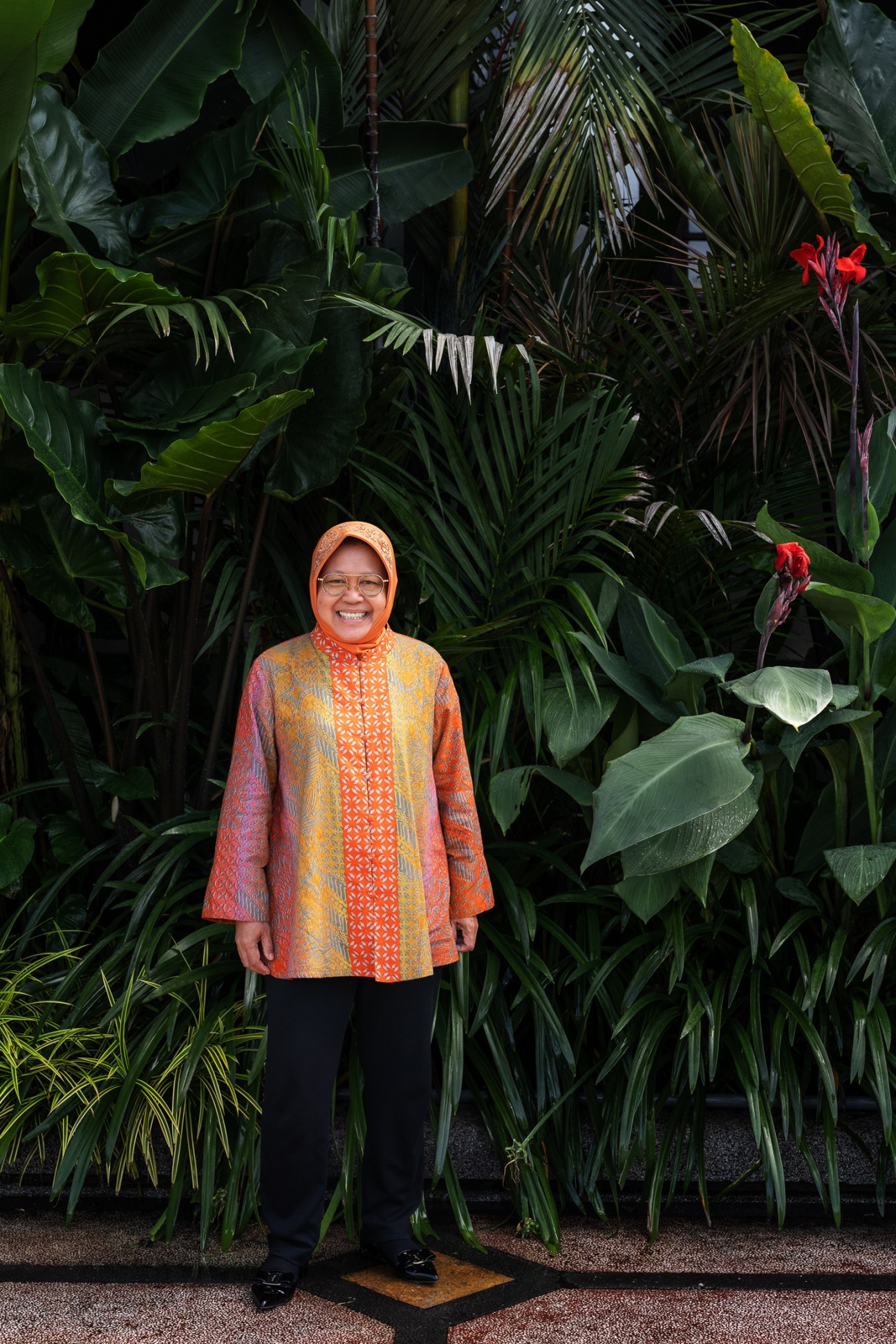Tri Rismaharini, Mayor of Surabaya, wearing orange headscarf and patterned jacket, standing in front of lush tropical plants