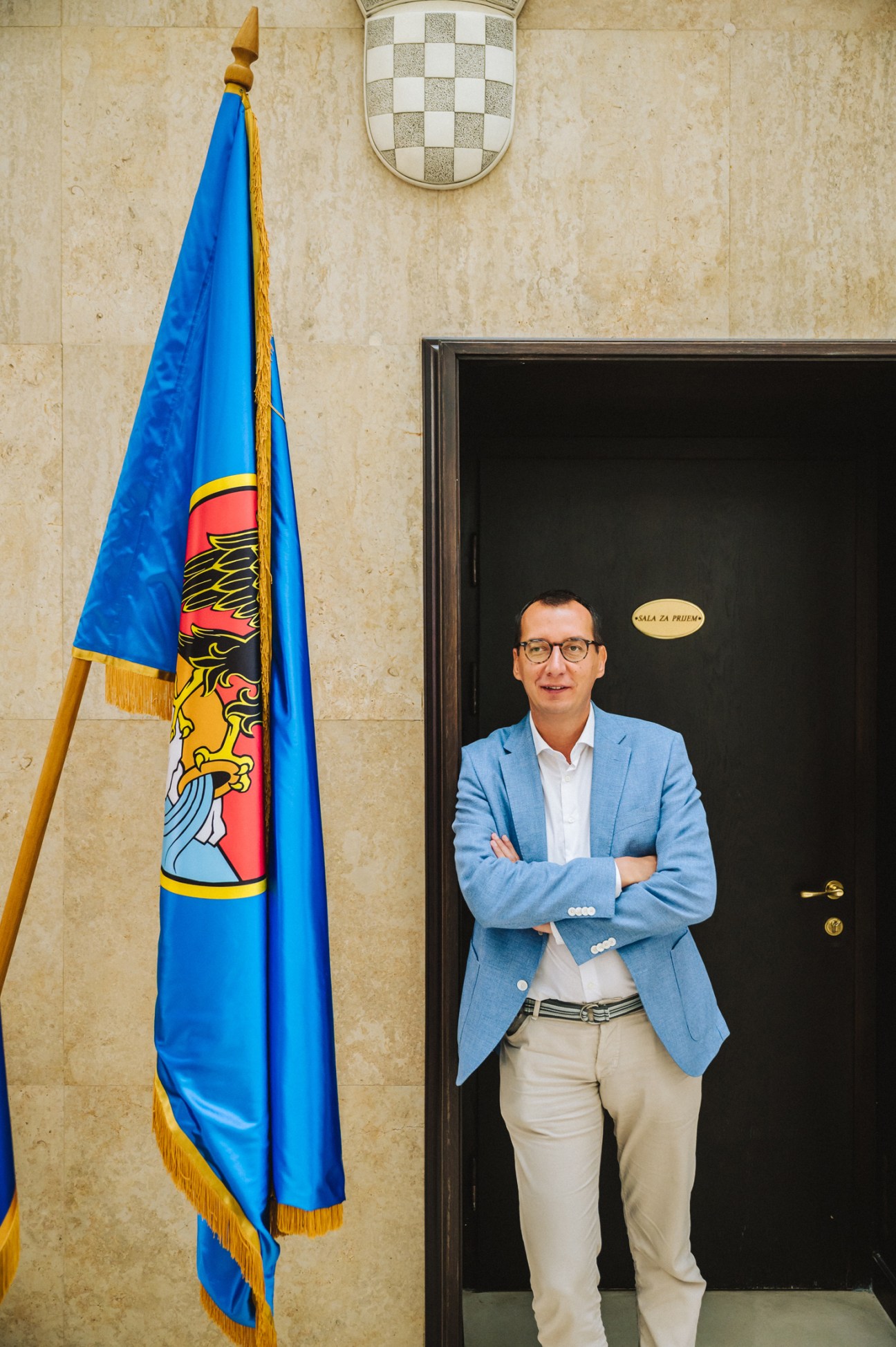 Mayor Marko Filipovic in a light blue blazer standing with crossed arms in front of a black door with coat of arms