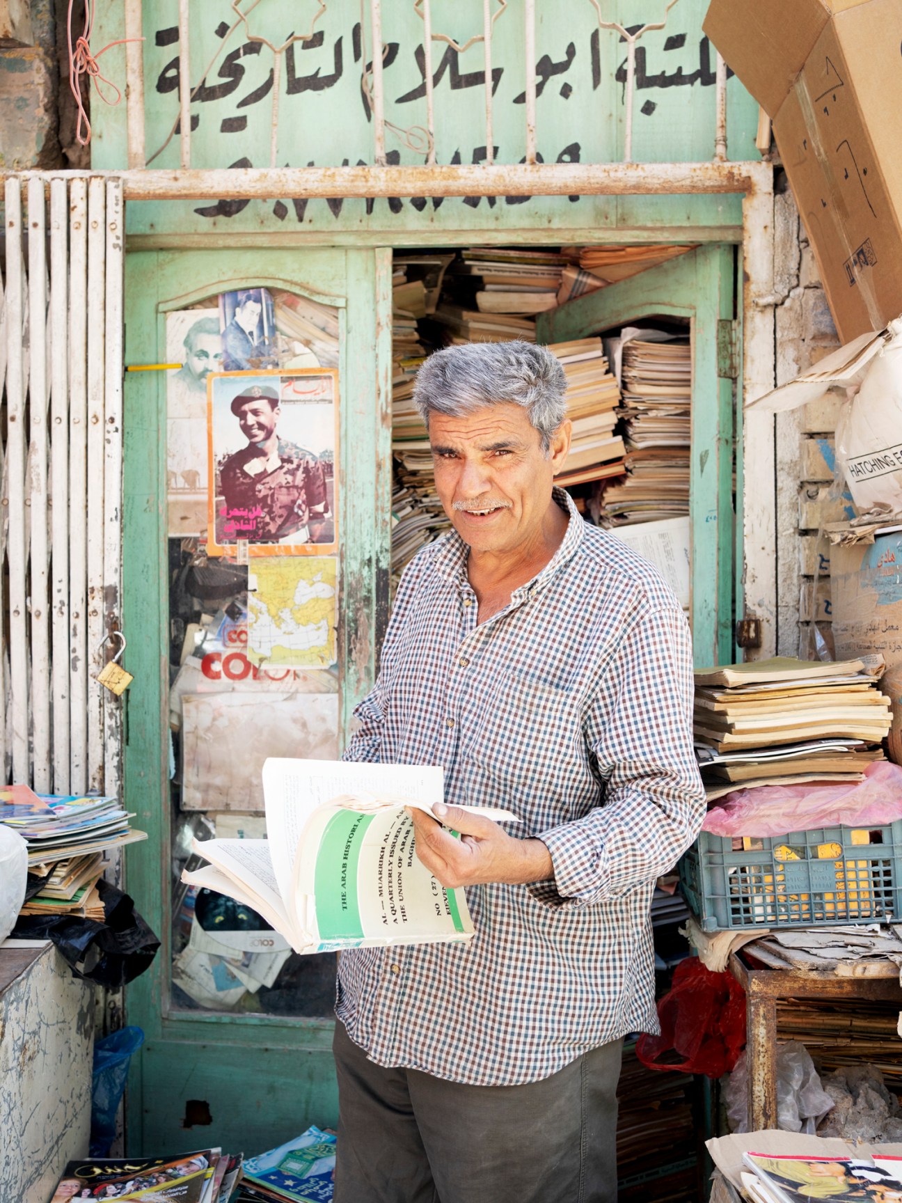 Man in checkered shirt holding book at Al Mutanabbi book market in Baghdad with stacks of books and Arabic signage