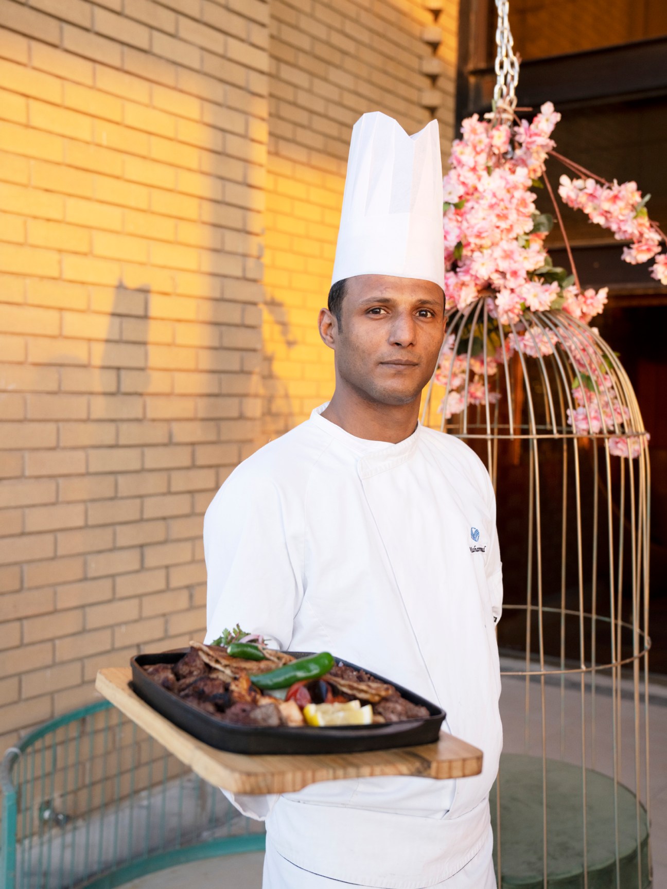 Chef in white uniform and hat holding a black serving platter with grilled meat and vegetables