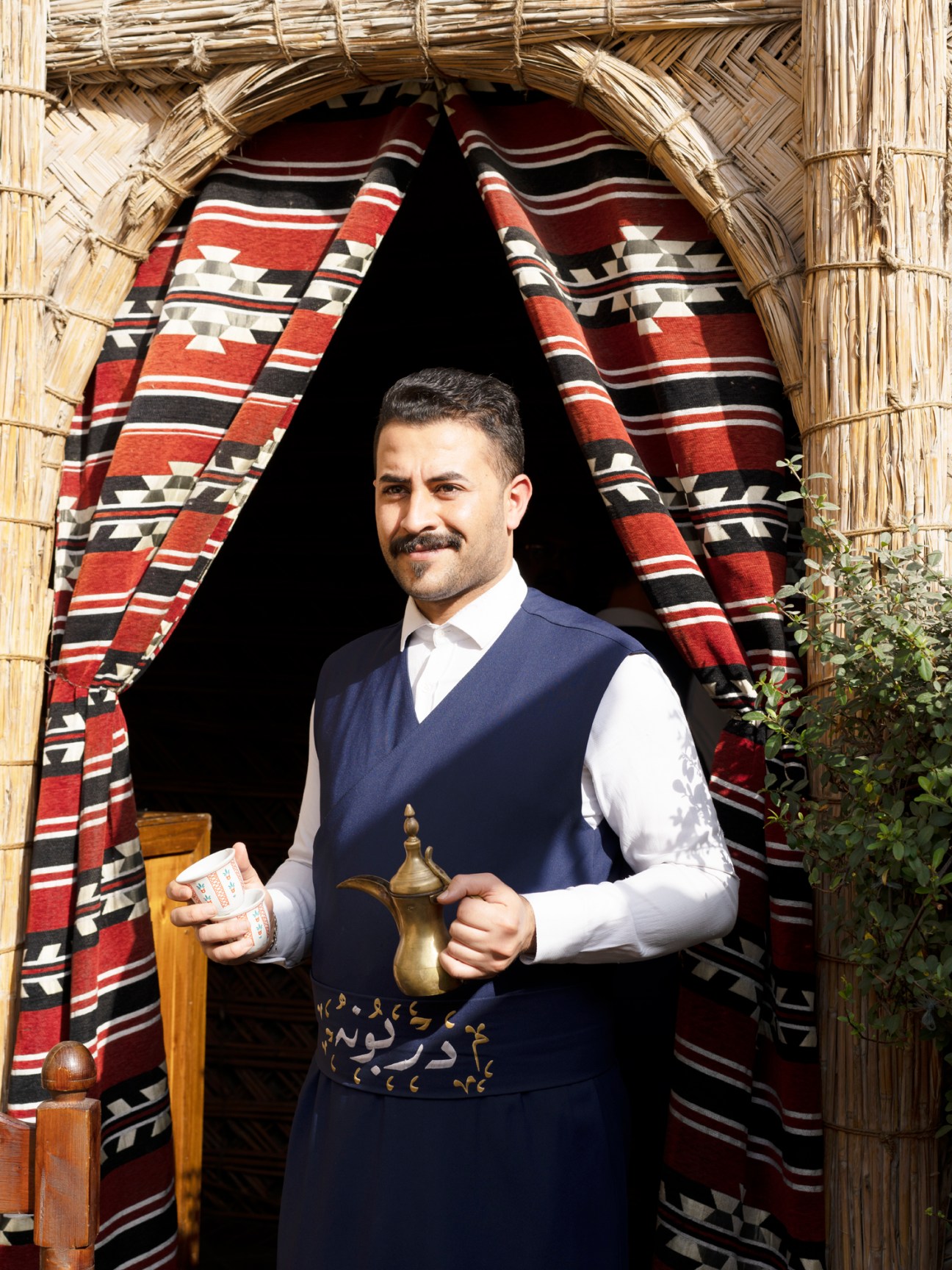 Man in traditional vest holding brass coffee pot at Darbunah restaurant in Baghdad