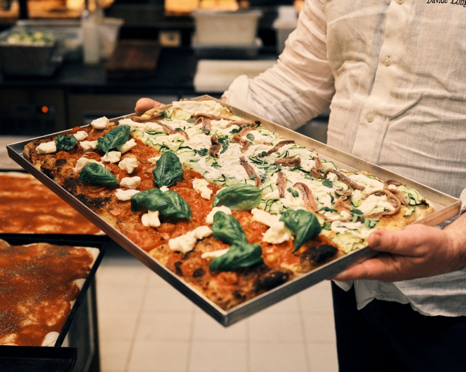 Person in striped chef's jacket holding a large rectangular pizza on a metal tray