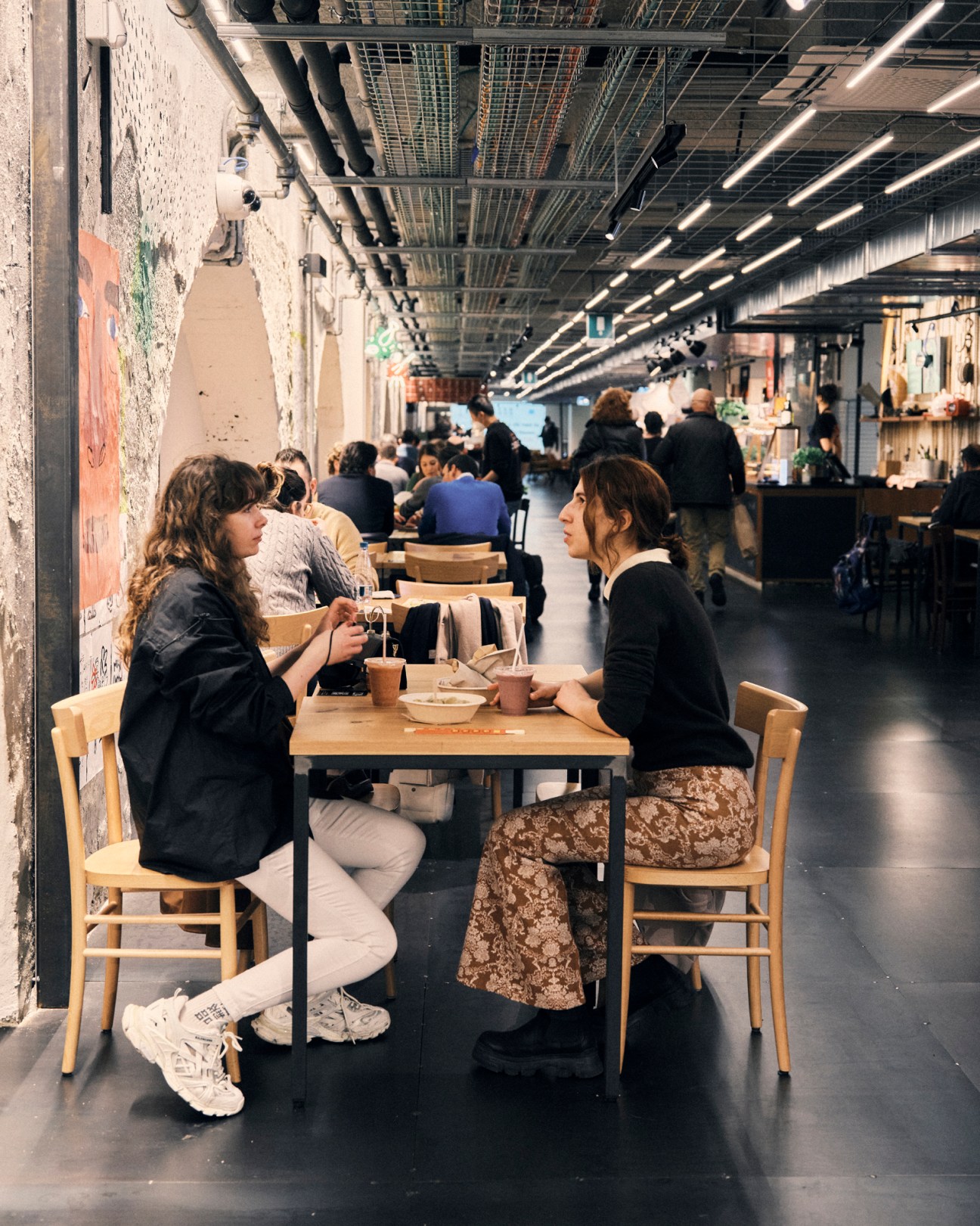 Two women sitting at a wooden table having coffee in a modern industrial-style dining area with exposed ceiling