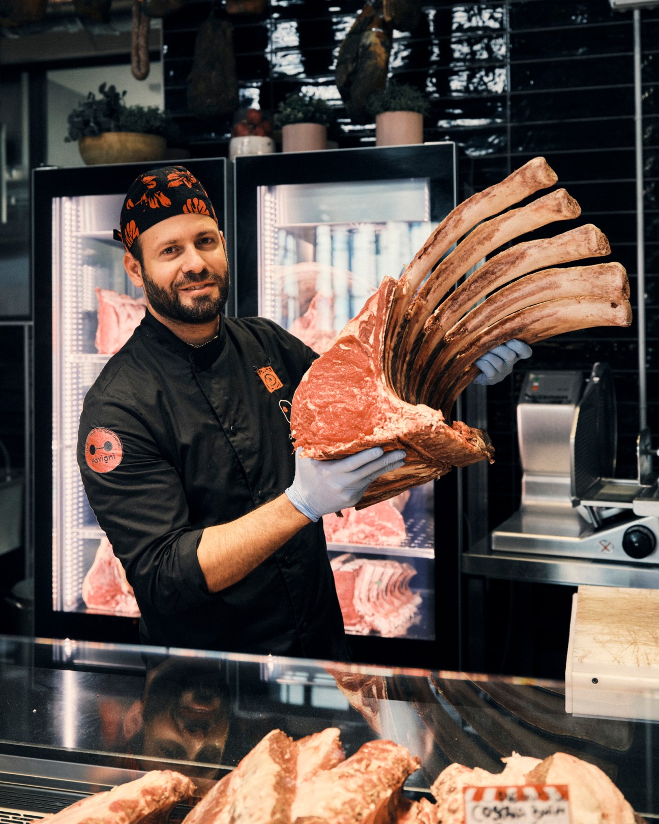 Butcher in black uniform and cap displaying large rack of ribs with long bones at meat counter