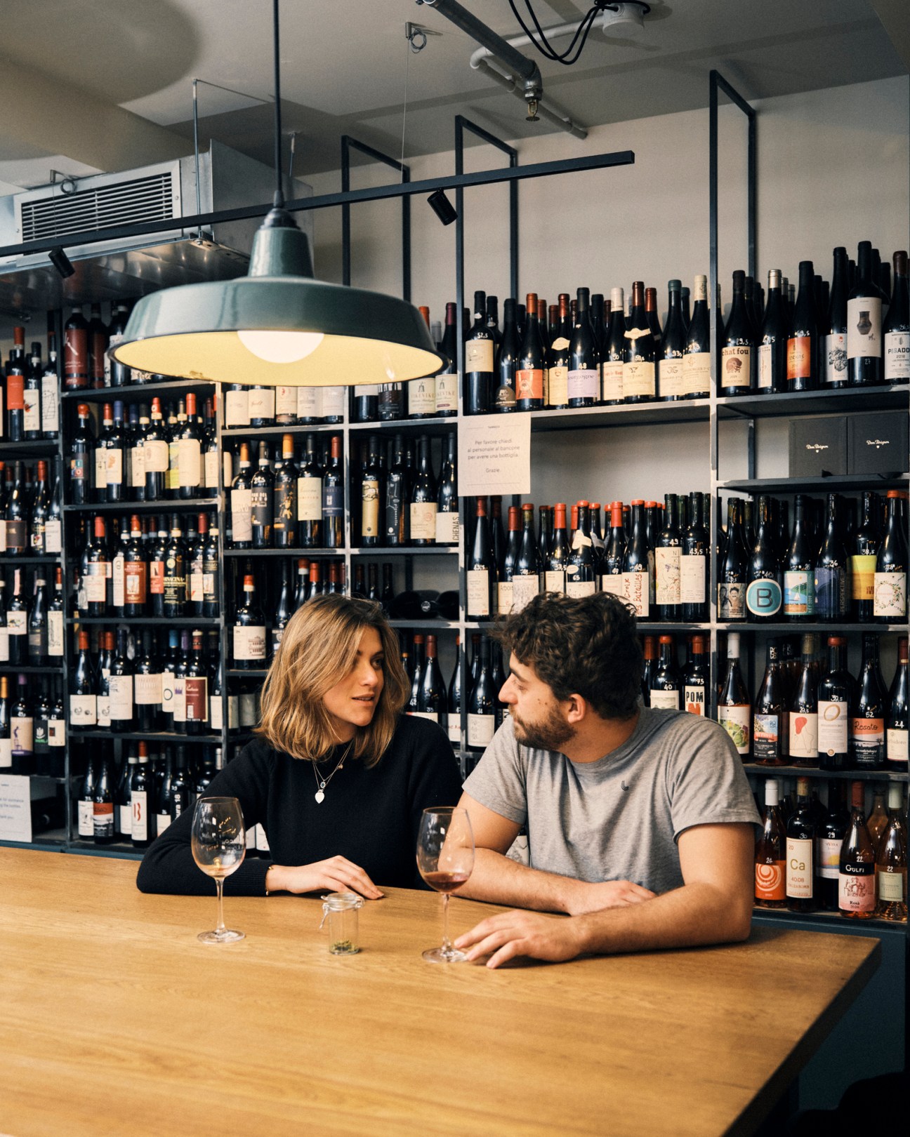 Two people sitting at a wooden counter in a wine shop with bottles lining the walls behind them