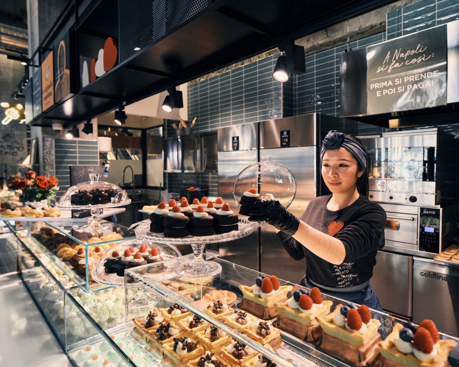 A pastry chef in black uniform and headband arranges desserts in a glass display case at a bakery