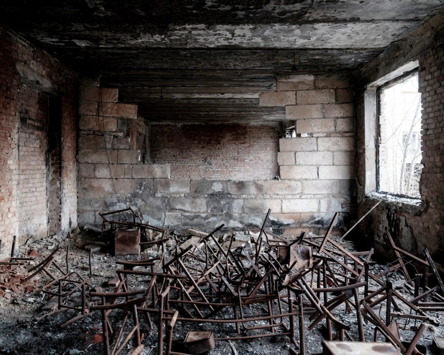 Destroyed classroom interior with collapsed ceiling, damaged brick walls, scattered broken chairs and debris